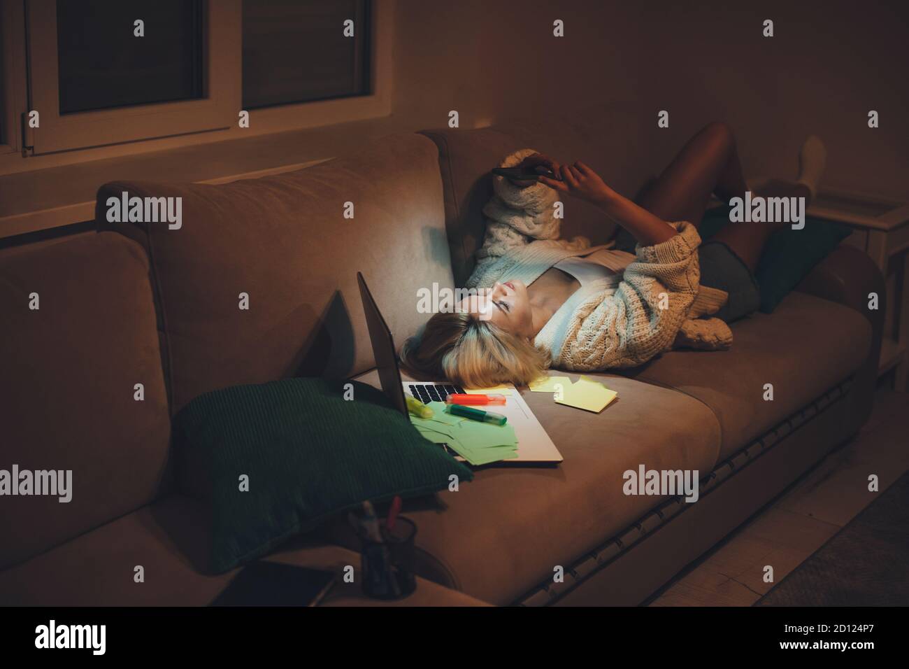 Donna caucasica mentita in letto che chiacchiera al telefono tardi a. notte dopo aver fatto i compiti con un computer portatile Foto Stock