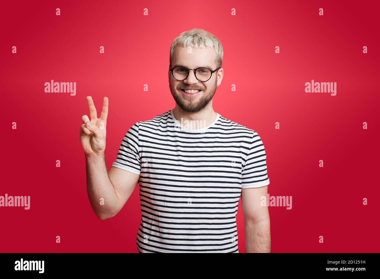 L'uomo caucasico con i capelli biondi e la barba è gesturing il cartello di pace su una parete rossa che sorride alla fotocamera Foto Stock