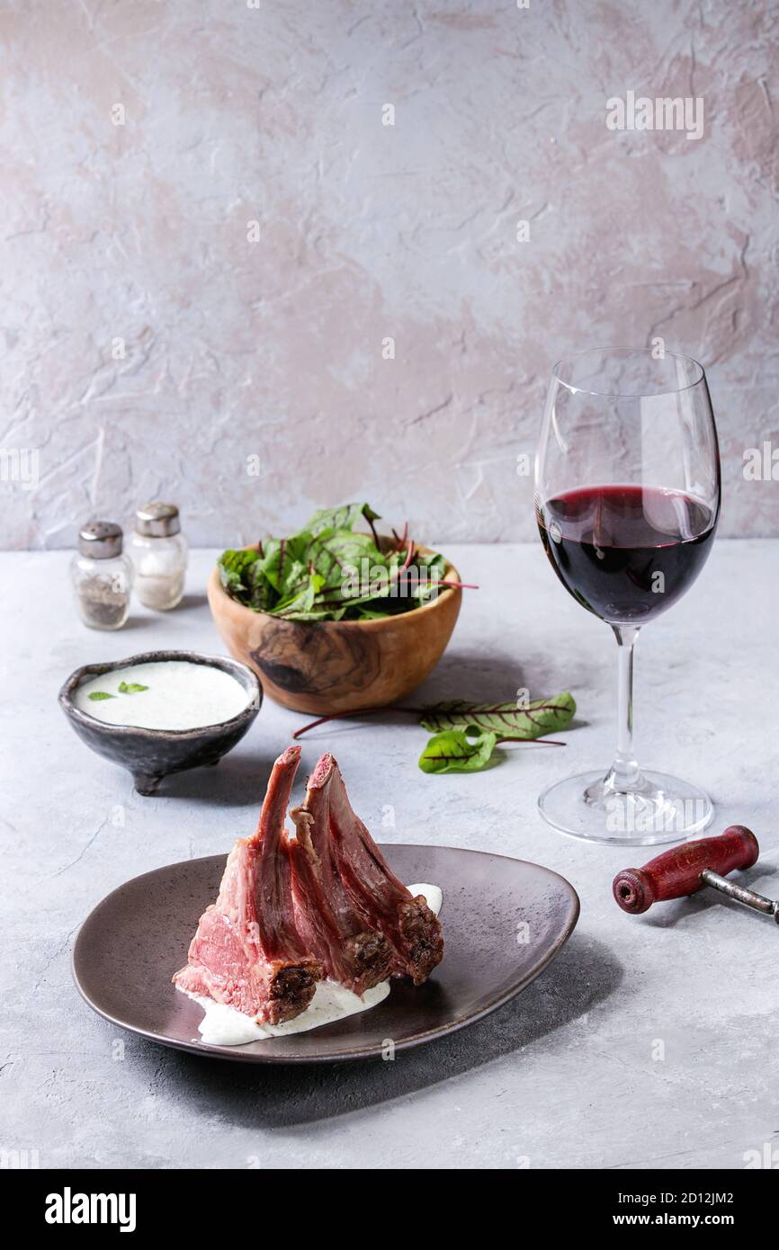 A fette grigliate carré di agnello con yogurt salsa alla menta servito nella piastra in ceramica con insalata verde giovani foglie di bietola, un bicchiere di vino rosso sulla texture grigio Foto Stock