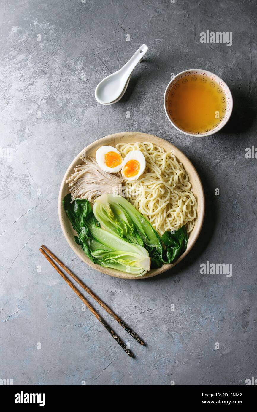 Ingredienti per la cottura piatto asiatico udon Tagliatelle con uova sode, funghi, boc choy, brodo servito in tazza ceramica con cucchiaio e bastoncini su grigio Foto Stock