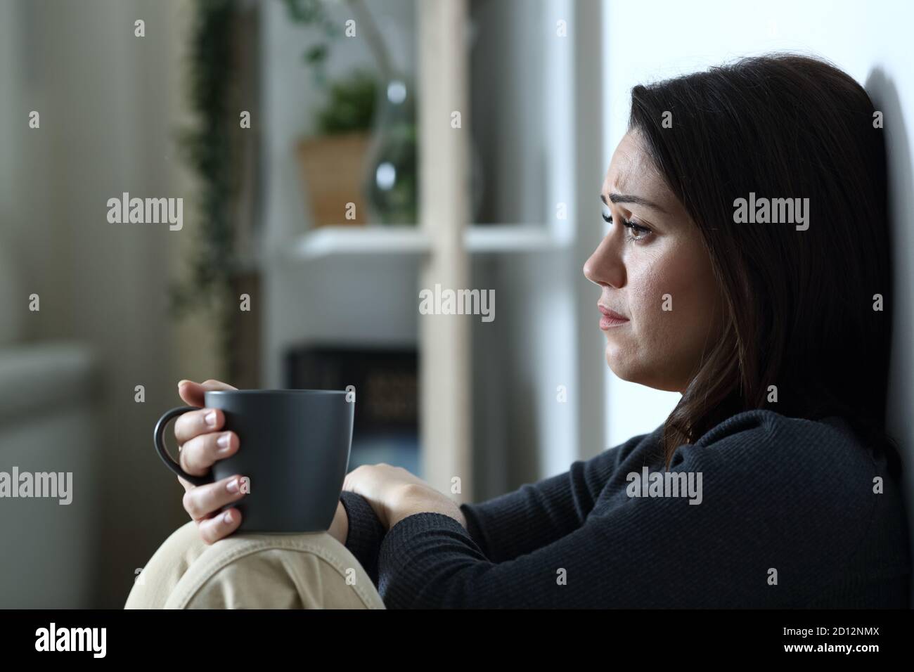 La donna triste con la tazza del caffè guarda via da sola a casa di notte Foto Stock