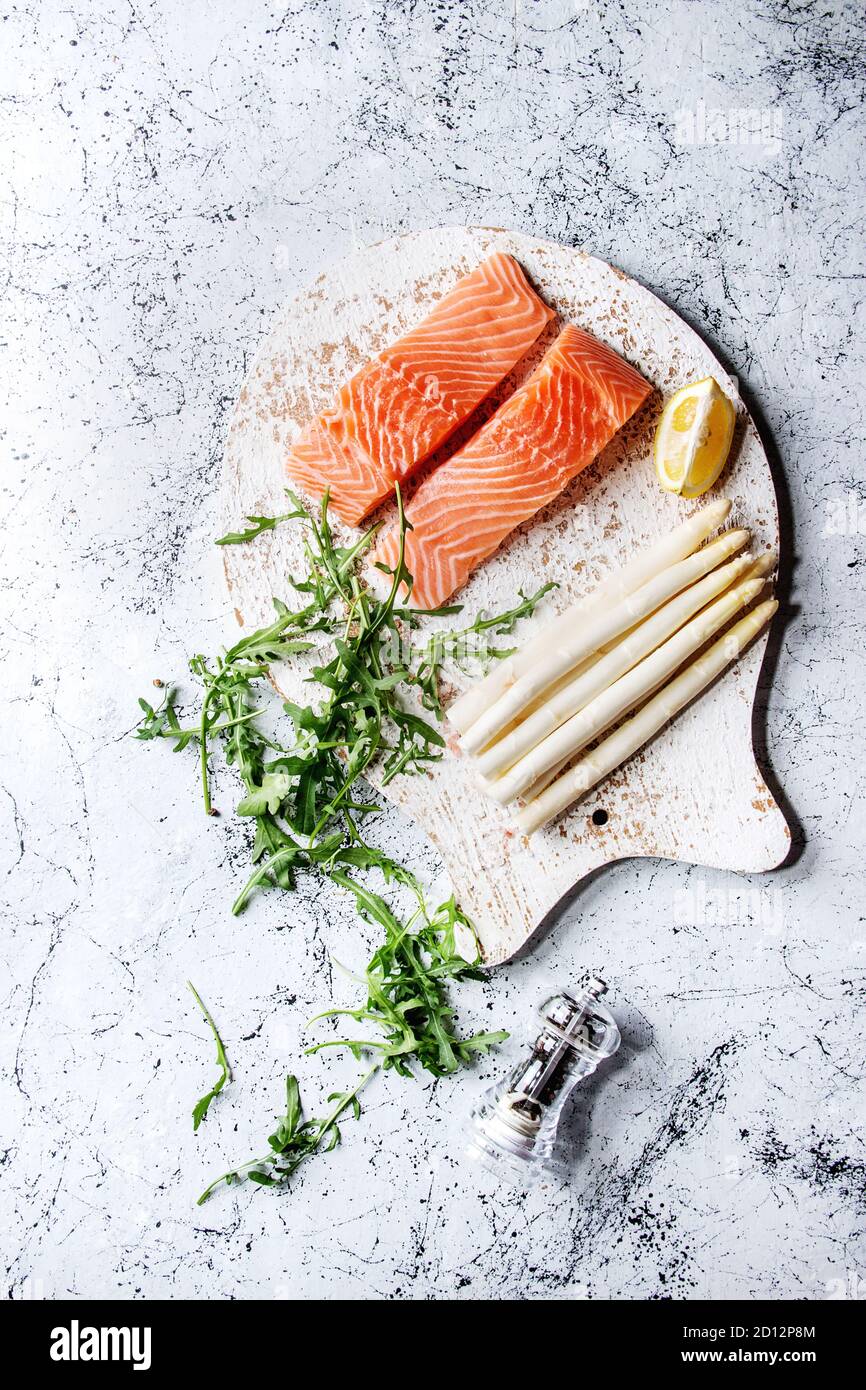 Fette di materie non cotte di filetto di salmone sul tagliere di legno a forma di pesce con gli ingredienti per cucinare la cena rucola, asparago bianco su bianco marbl Foto Stock