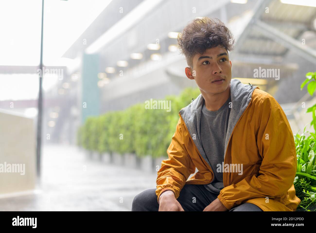 Giovane uomo asiatico con capelli ricci pensando e seduto in la città all'aperto con la pioggia Foto Stock