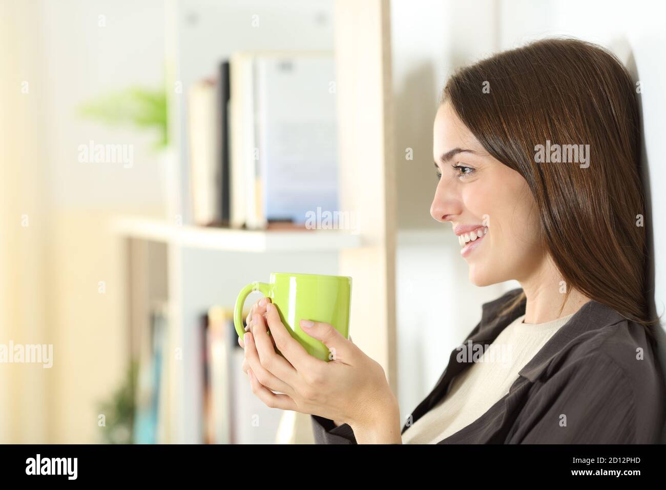 Vista laterale ritratto di una donna felice che tiene una tazza di caffè guarda lontano in piedi a casa Foto Stock