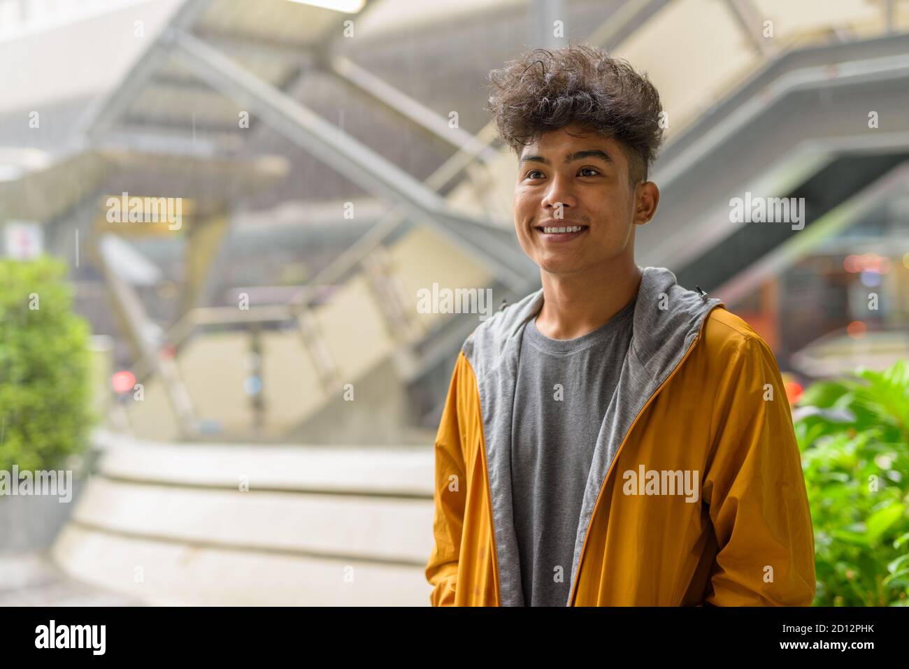 Felice giovane uomo asiatico con capelli ricci pensando nel città all'aperto con la pioggia Foto Stock