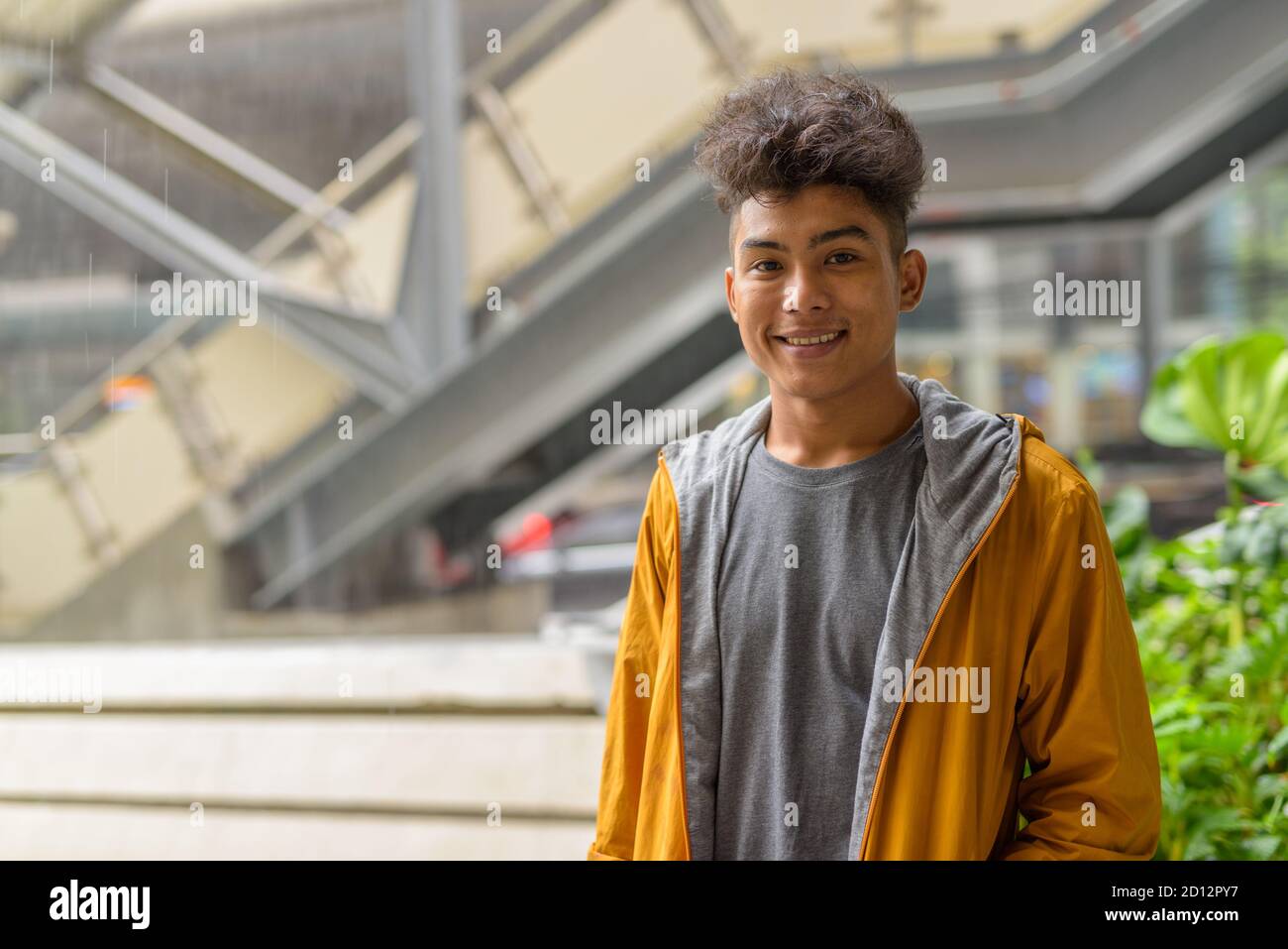 Felice giovane asiatico con capelli ricci sorridenti nel città all'aperto con la pioggia Foto Stock