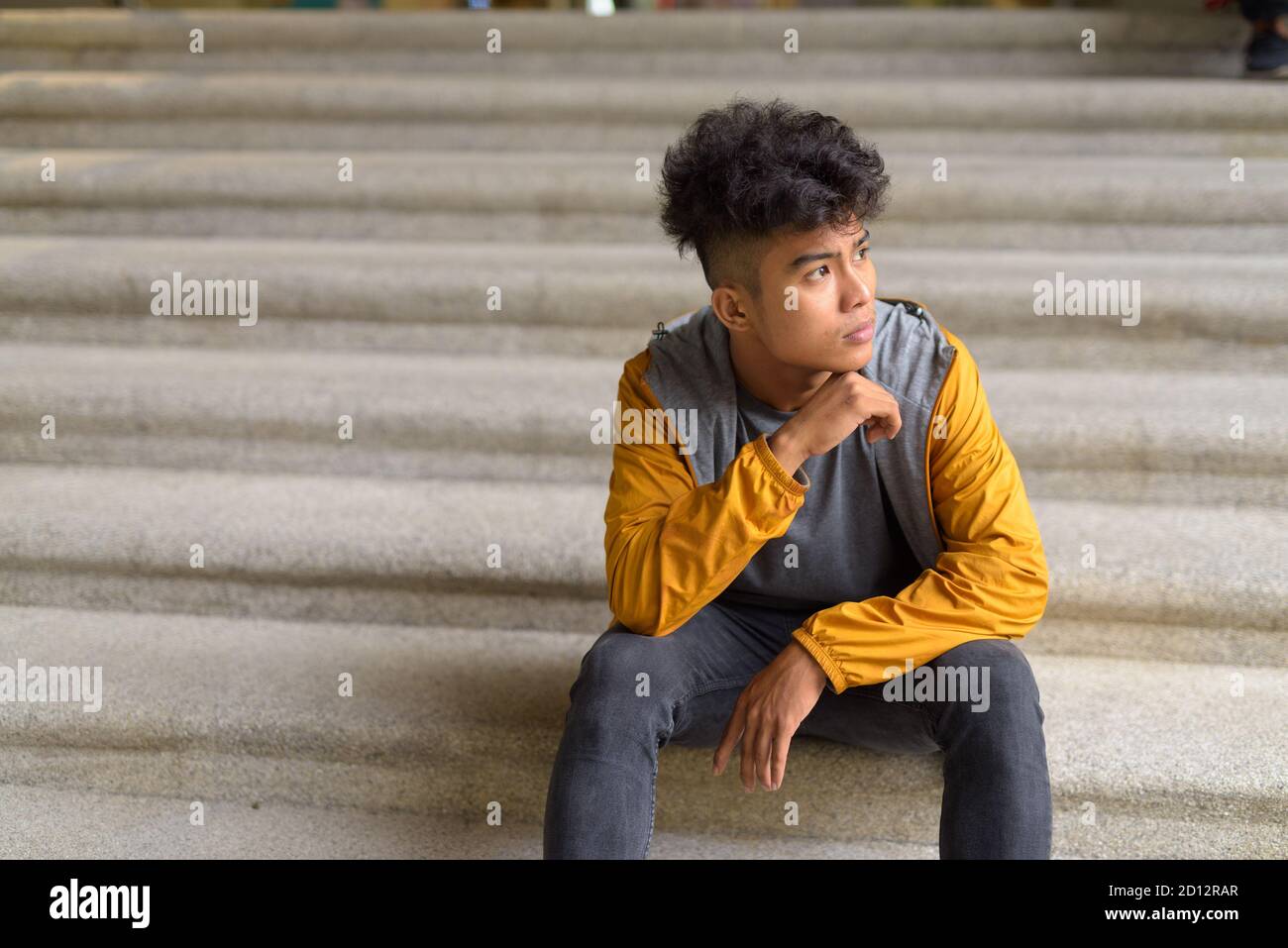 Giovane uomo asiatico con capelli ricci che pensano e si siede sopra scala in città Foto Stock