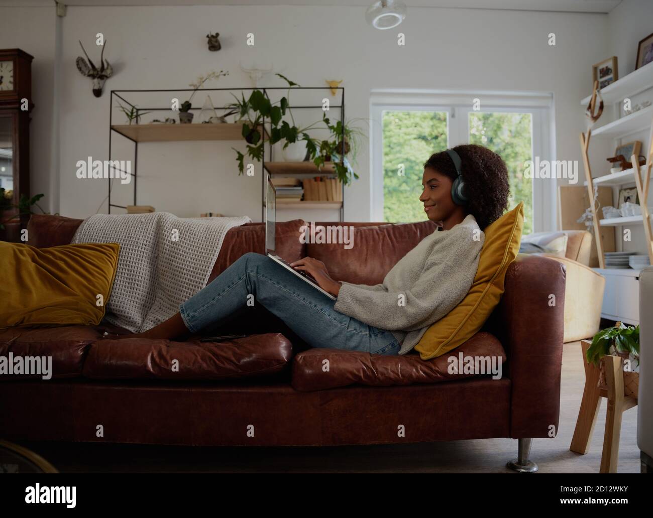Vista laterale di una giovane donna che si rilassa a casa guardando il webinar e digitando con le cuffie seduti sul divano Foto Stock