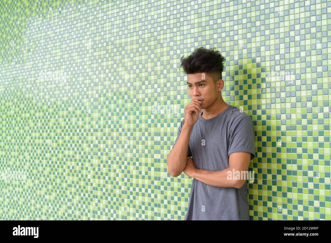 Giovane uomo asiatico con capelli ricci che pensano e si appoggia contro parete verde all'aperto Foto Stock