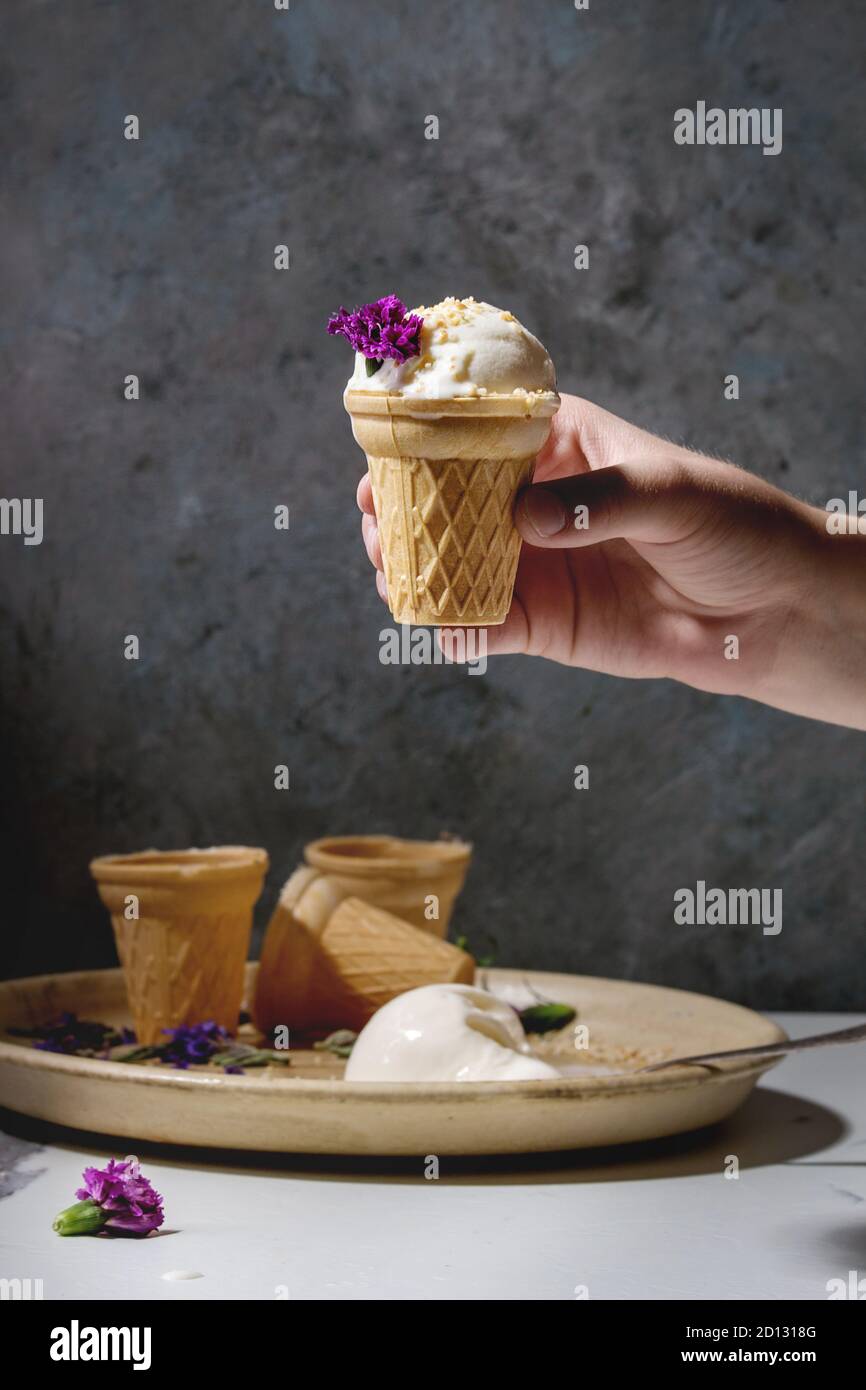 Bambino la mano a prendere in casa di gelato alla vaniglia in cialda di piccola tazza servita con viola fiori commestibili e cucchiaio di metallo nella piastra in ceramica su marmo bianco ki Foto Stock