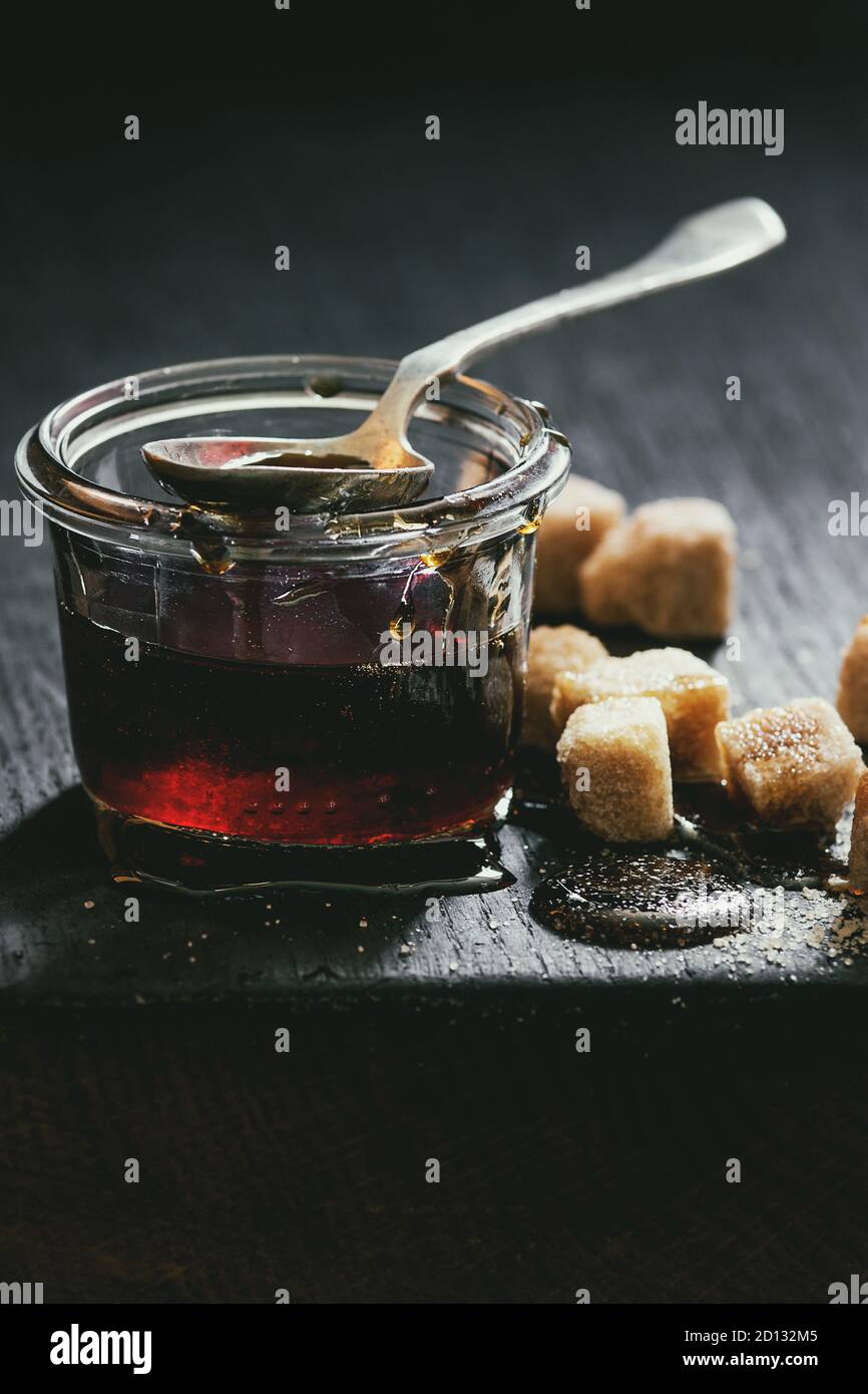 In casa liquido trasparente marrone zucchero caramellato in un barattolo di vetro in piedi sul legno nero con bordo cucchiaio e può zollette di zucchero. Close up. La luce del giorno. Foto Stock