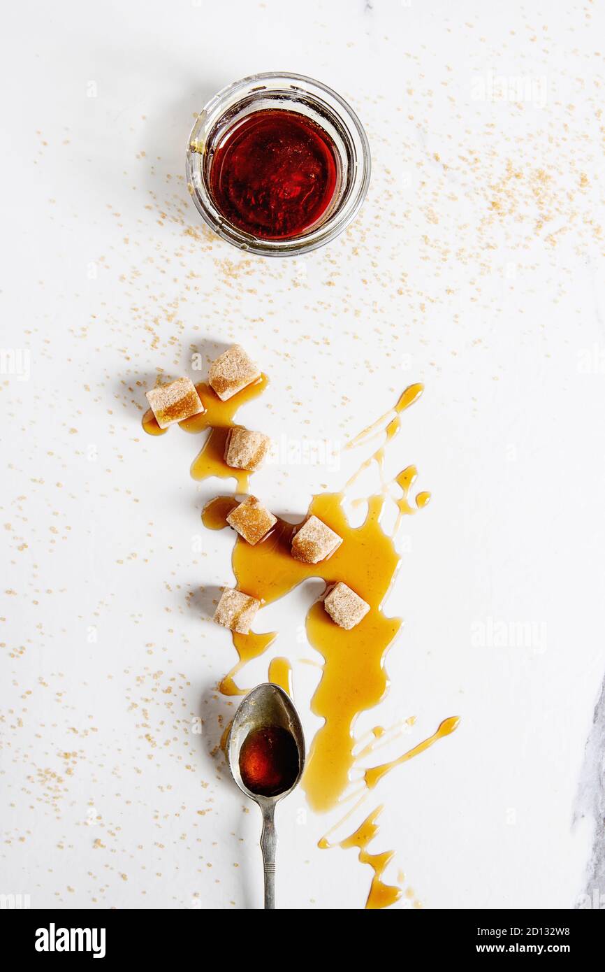 In casa liquido trasparente marrone zucchero caramellato in un barattolo di vetro con il cucchiaio e la canna da zucchero Zollette di zucchero bianco su sfondo marmo. Appartamento laico, spazio. Foto Stock