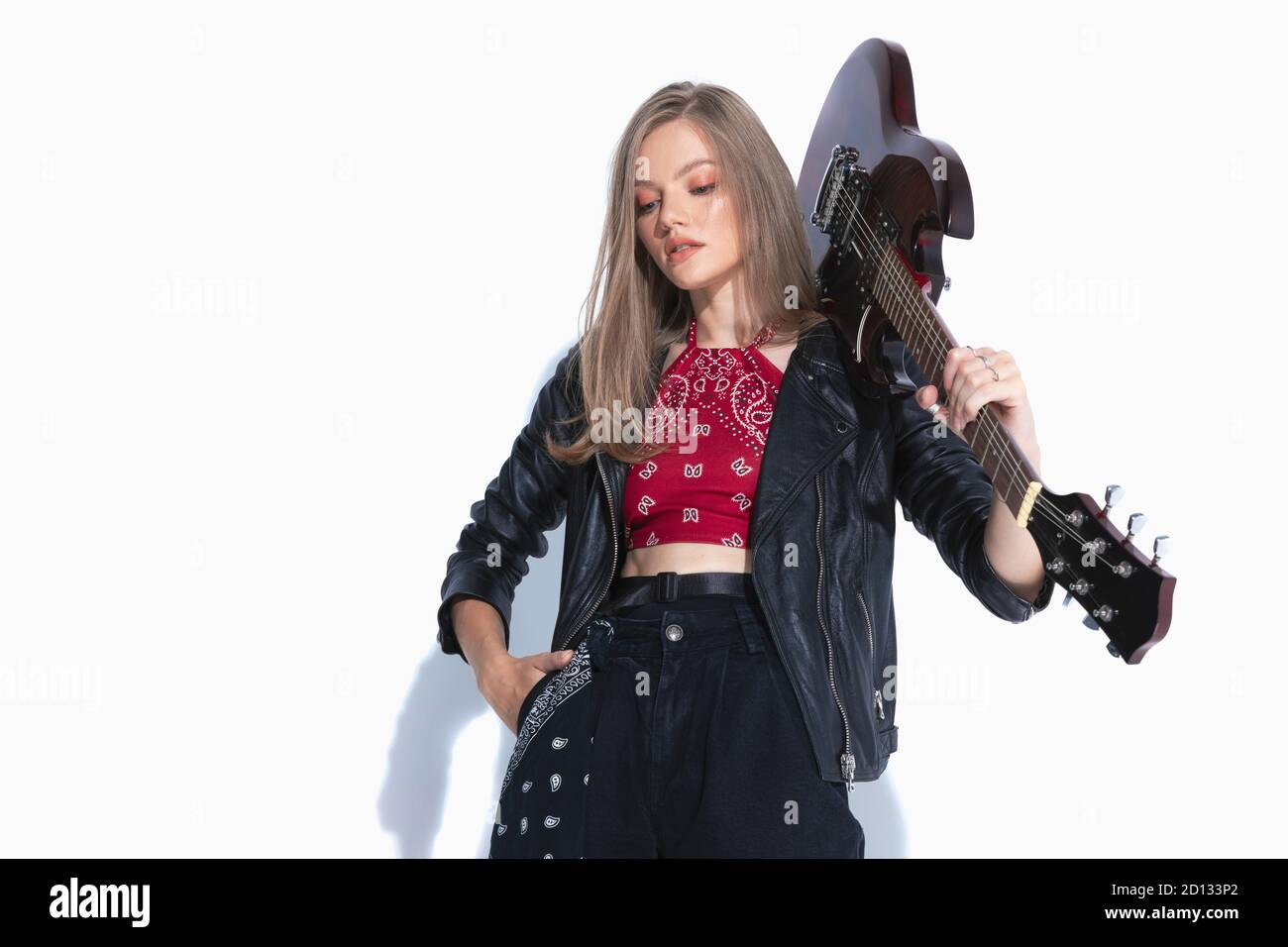 Un musicista determinato che tiene la chitarra sulla spalla e la mano in tasca, indossando una giacca in pelle mentre si trova su uno sfondo bianco dello studio Foto Stock