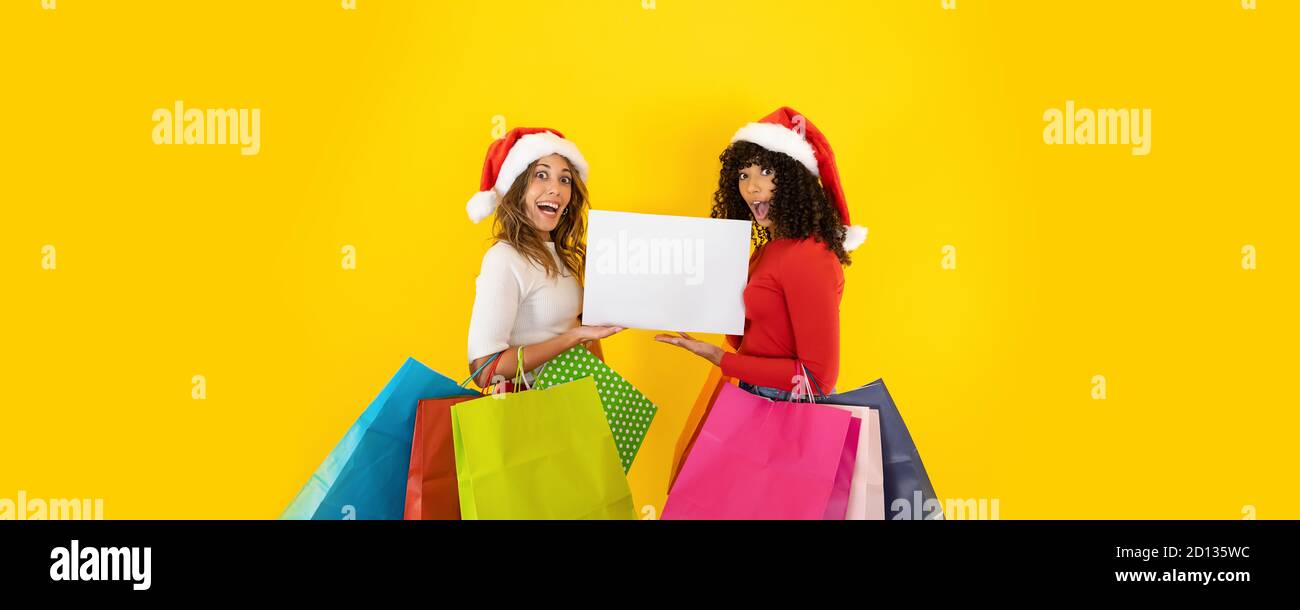 Due giovani donne carine che indossano il cappello di Babbo Natale guardando alla macchina fotografica con viso stupefatto, bocca aperta, occhi larghi, tenendo borse colorate per la spesa - Cristo Foto Stock