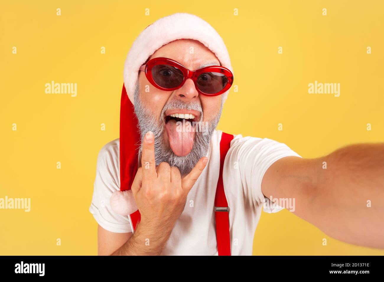 Uomo pazzo estremamente eccitato nel cappello di babbo natale e occhiali da sole che imbrogliano le facce guardando la macchina fotografica, blogger anziani che si divertono mostrando gesto rock. Interno Foto Stock