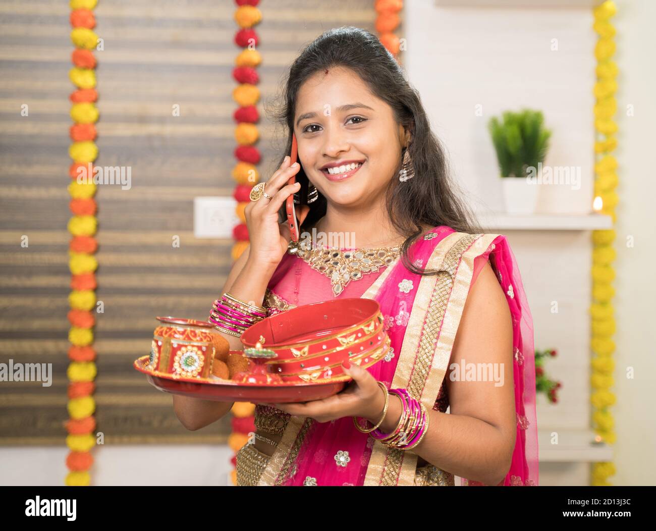 Sorridente donna indiana in abito tradizionale che tiene Karva Chauth Thali o piatto mentre occupato a parlare sul cellulare. Foto Stock