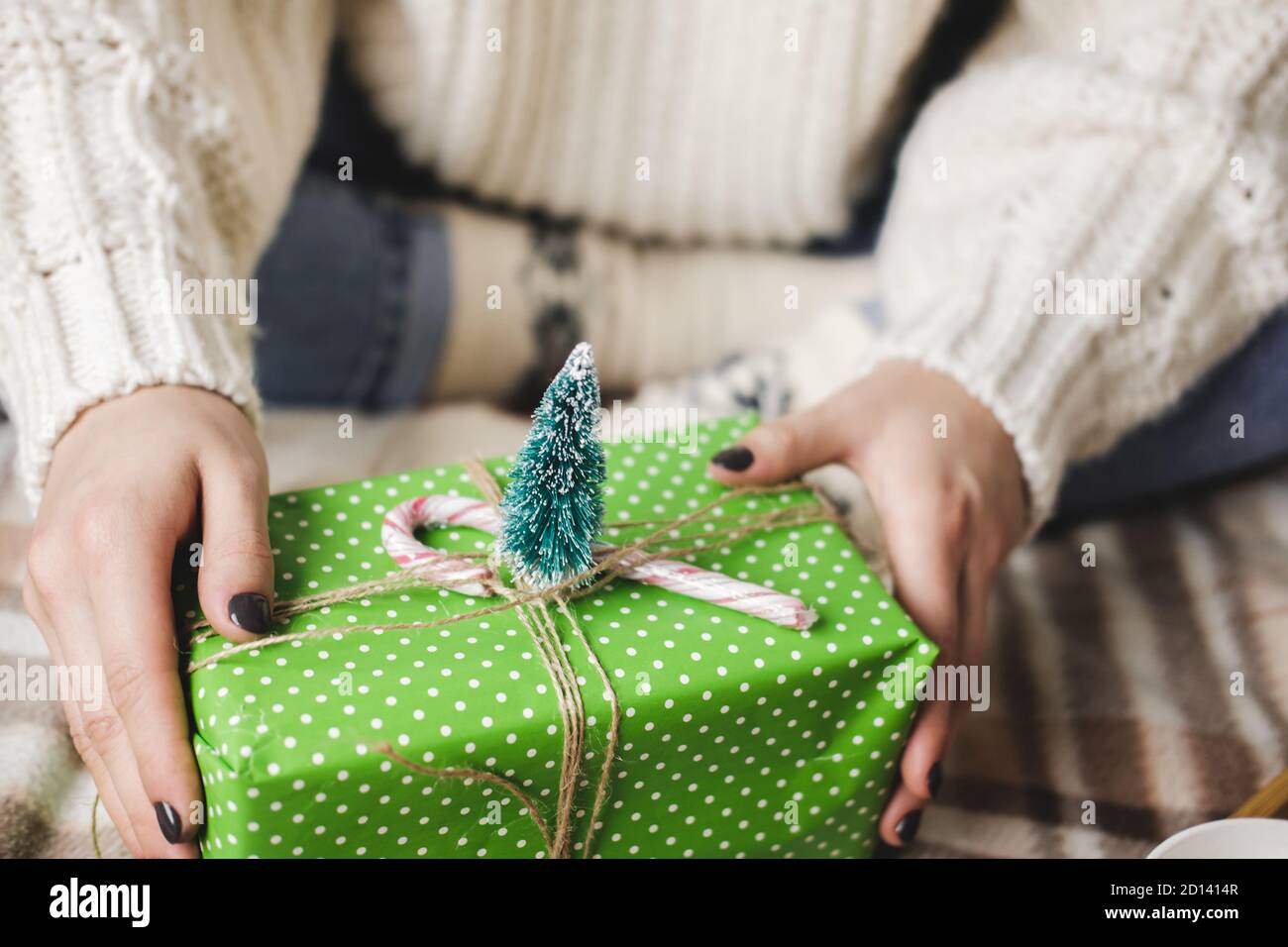 Giovane donna si siede su plaid in maglia accogliente pullover bianco lana, calzini e avvolge regalo di Natale in polka dot wrapping carta. Vassoio in legno con tazza di co Foto Stock