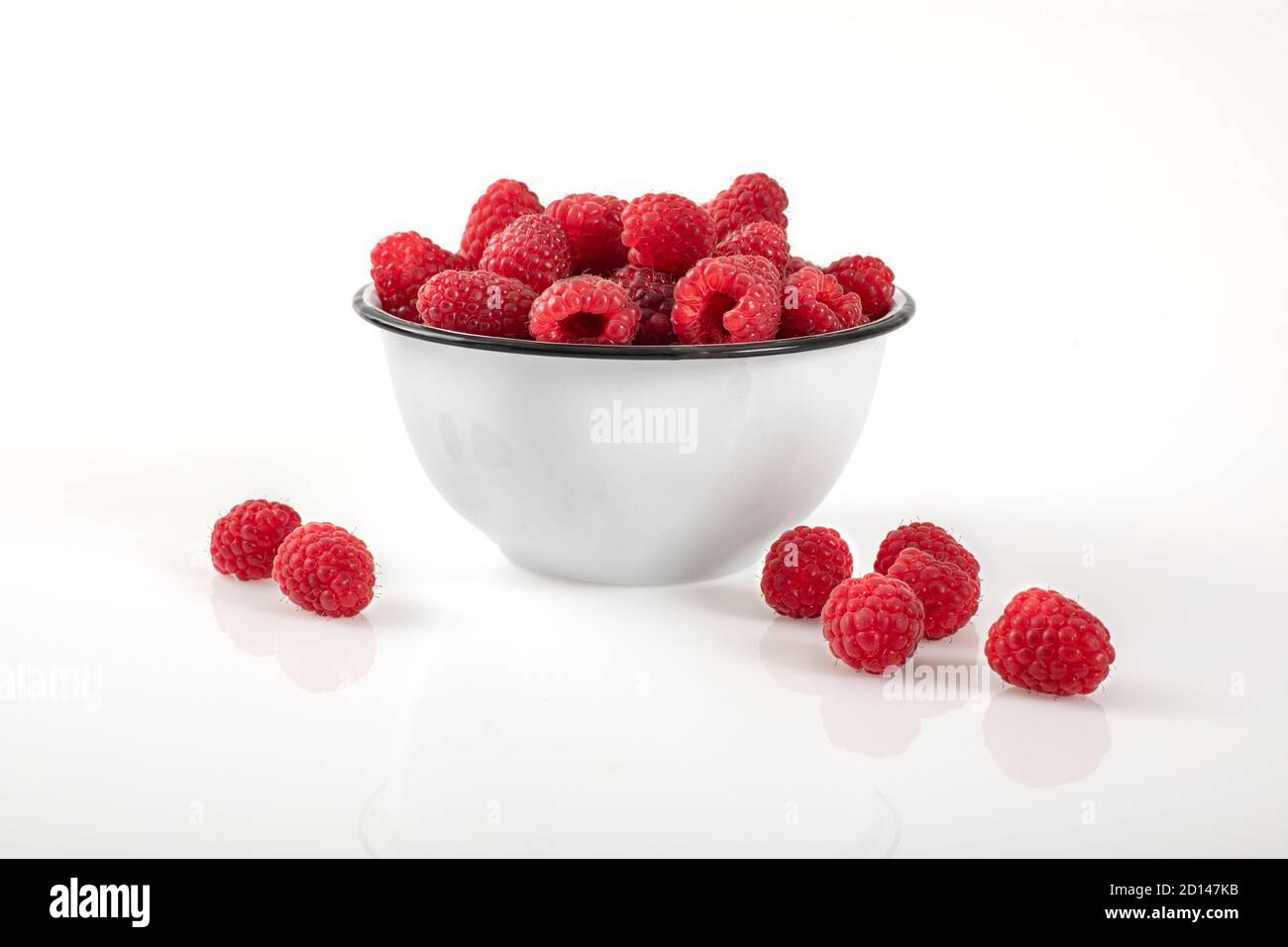 Un sacco di lamponi in un recipiente posto su sfondo bianco e lucido. Alcuni frutti di bosco sono posti sulla superficie esterna al recipiente. Foto Stock