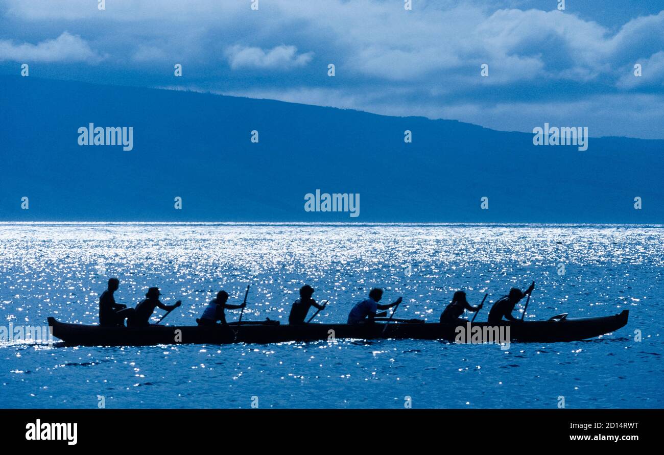 Una squadra di sei persone che si allenano per una corsa in una tradizionale canoa hawaiana Outrigger è silhouette dalla luce solare che si affaccia sull'Oceano Pacifico settentrionale al largo della costa di Maui, nelle Hawaii, USA. Le tipiche canoe da corsa hawaiane hanno un singolo grilletto su un lato dell'imbarcazione che fornisce stabilità alla lunga ed elegante barca. Tre membri dell'equipaggio tirano attraverso l'acqua su ogni lato con pale che sono dotate di manici lunghi con una singola lama ad un'estremità. Anche visto qui è un settimo uomo, un allenatore che si siede più in alto nella poppa (posteriore) della barca durante la pratica, ma non è mai a bordo durante le gare reali. Foto Stock