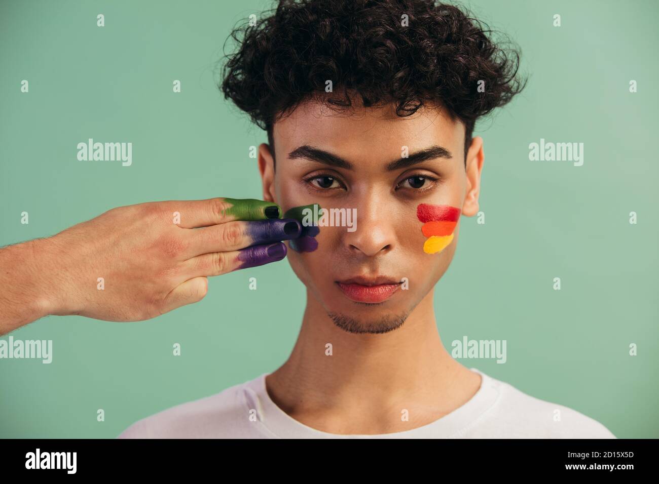 Closeup di una mano pittura LGTB bandiera colori sul viso dell'uomo. Uomo transgender con bandiera gay di orgoglio dipinta sulle sue guance. Foto Stock