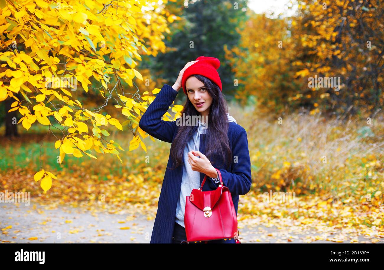 Bruna sorridente che corregge il suo cappello rosso nel parco autunnale durante l'estate indiana. Ritratto donna all'aperto nel parco Foto Stock