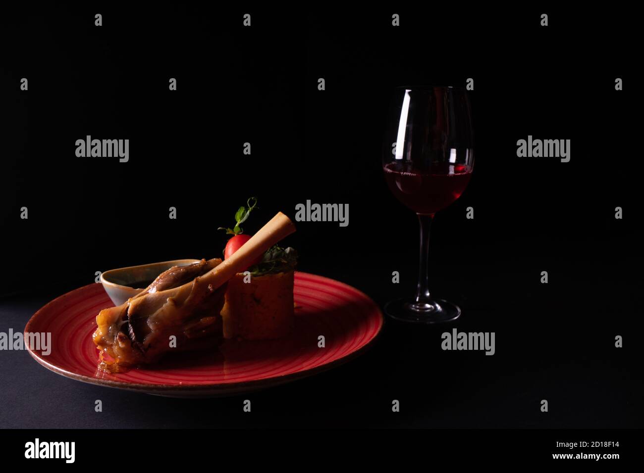 Gamba di agnello con salsa su un piatto rosso e un bicchiere di vino su sfondo nero Foto Stock