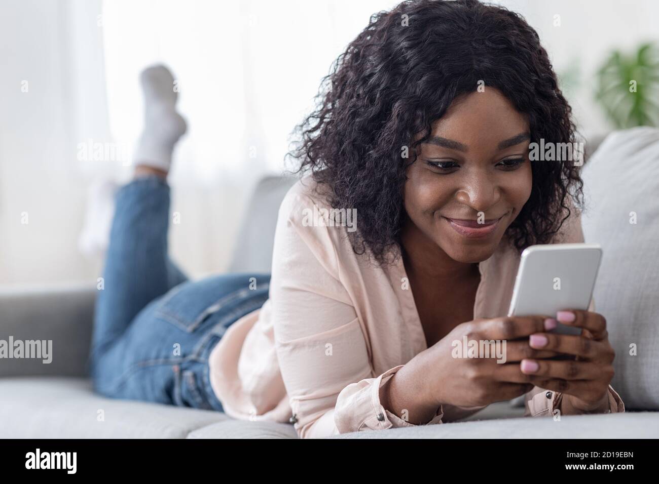 Sorridente donna africana passare il tempo con lo smartphone a casa, messaggi con gli amici Foto Stock
