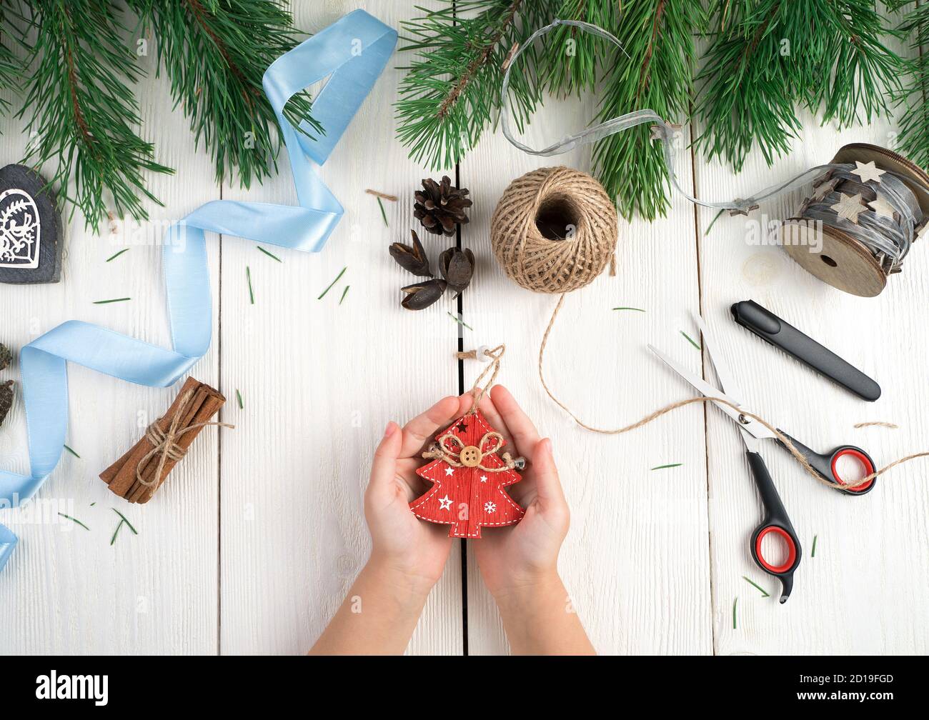 Albero di Natale in legno rosso nelle mani di un bambino, rami di abete, nastri, ingushki su uno sfondo di legno chiaro. Foto Stock