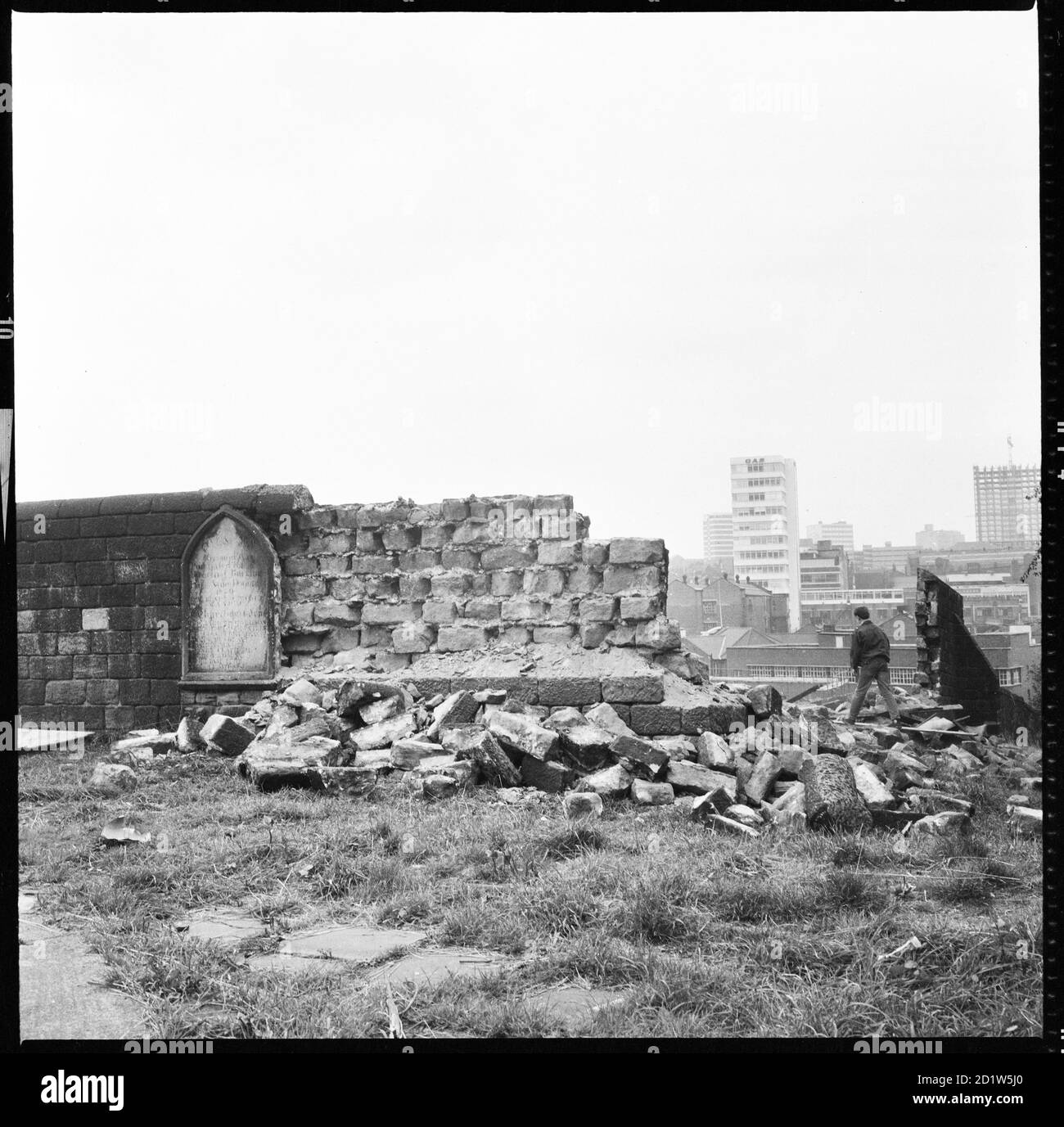 Un ragazzo che cammina attraverso la lapide demolita del muro del cimitero presso la chiesa di St Mary con gli edifici su Mabbate sullo sfondo e gli uffici moderni del centro della città in lontananza, St Mary's Street, Quarry Hill, Leeds, UK. Foto Stock