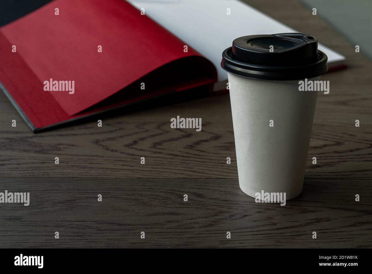 Caffè del mattino e un libro. Una tazza da caffè bianca usa e getta con coperchio nero su un tavolo di legno con libro aperto sullo sfondo. Horizontas Foto Stock