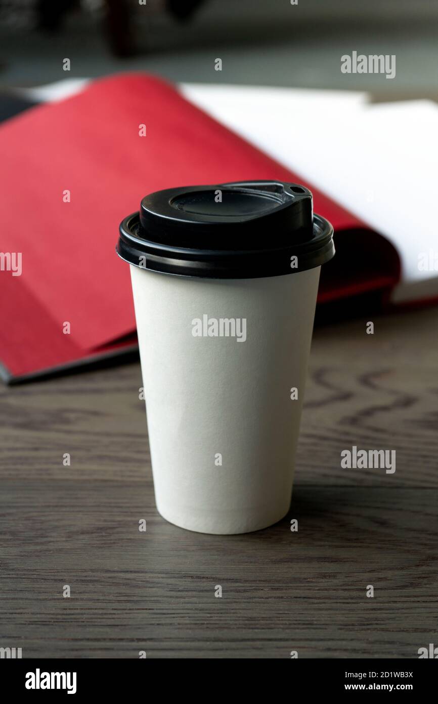 Caffè del mattino e un libro. Una tazza da caffè bianca usa e getta con coperchio nero su un tavolo di legno con libro aperto sullo sfondo. Verticale Foto Stock