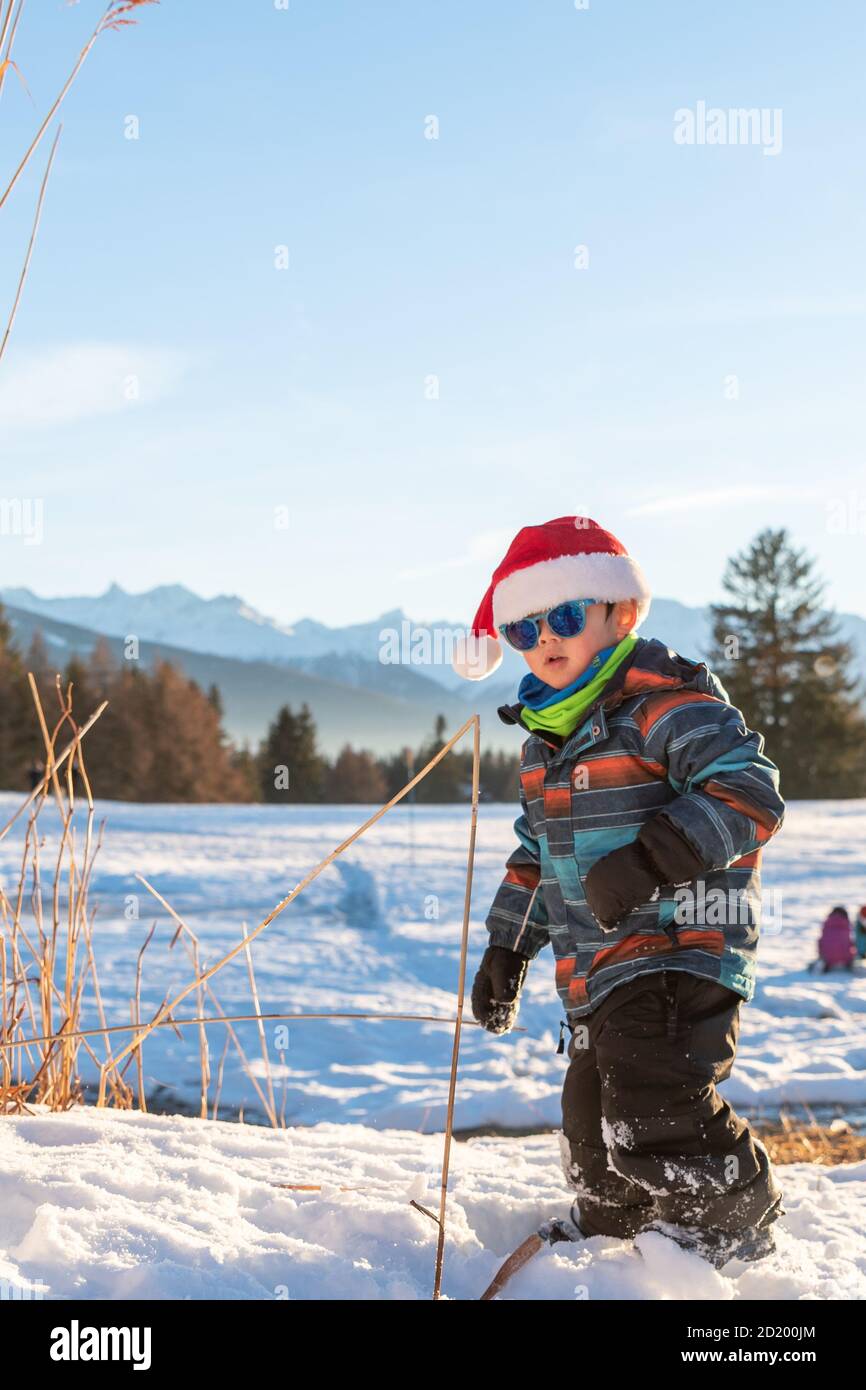 Ragazzo con cappello rosso Babbo Natale su uno sfondo di paesaggio invernale. Bambino sorridente felice nella neve a Natale in piedi accanto a piante alte all'aperto. Gru Foto Stock