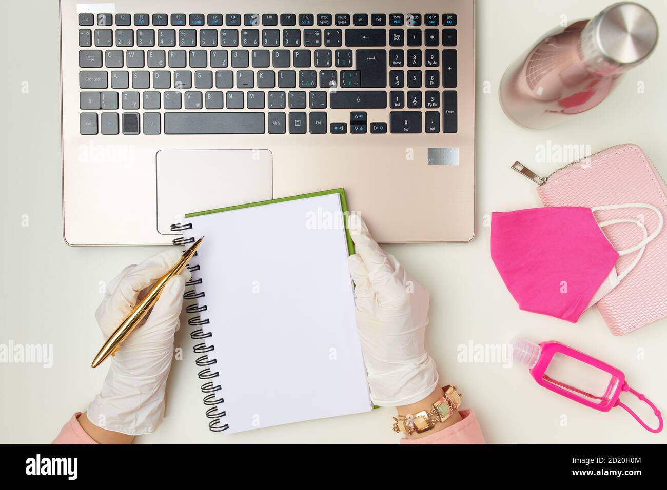 Vista superiore delle mani femminili in guanti di gomma bianca che scrivono in un taccuino vicino a maschera medica, antisettico e portafoglio su tavolo bianco. Foto Stock