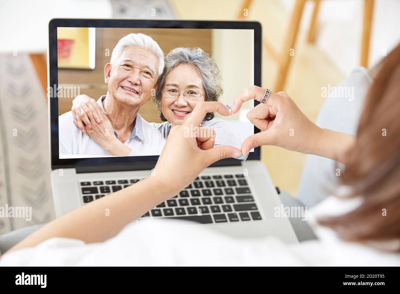 giovane figlia asiatica adulta che rimane a casa formando un cuore forma con le mani mentre parla con i genitori anziani in linea via chat video con computer portatile Foto Stock