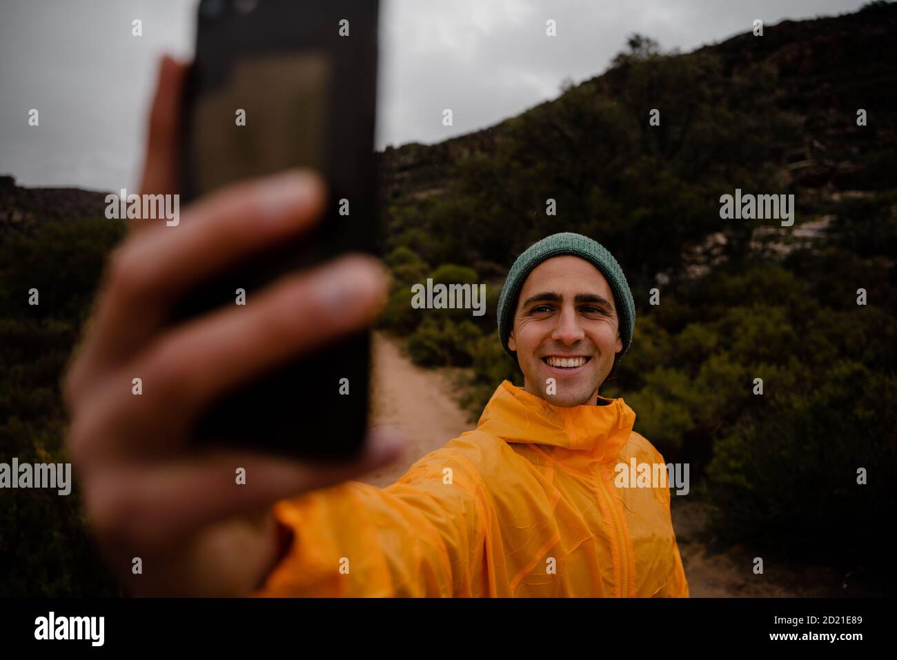 Bel giovane corridore di sentiero maschio sorridente scattando foto di sé su smartphone su sentiero di montagna in tempo freddo nuvoloso Foto Stock