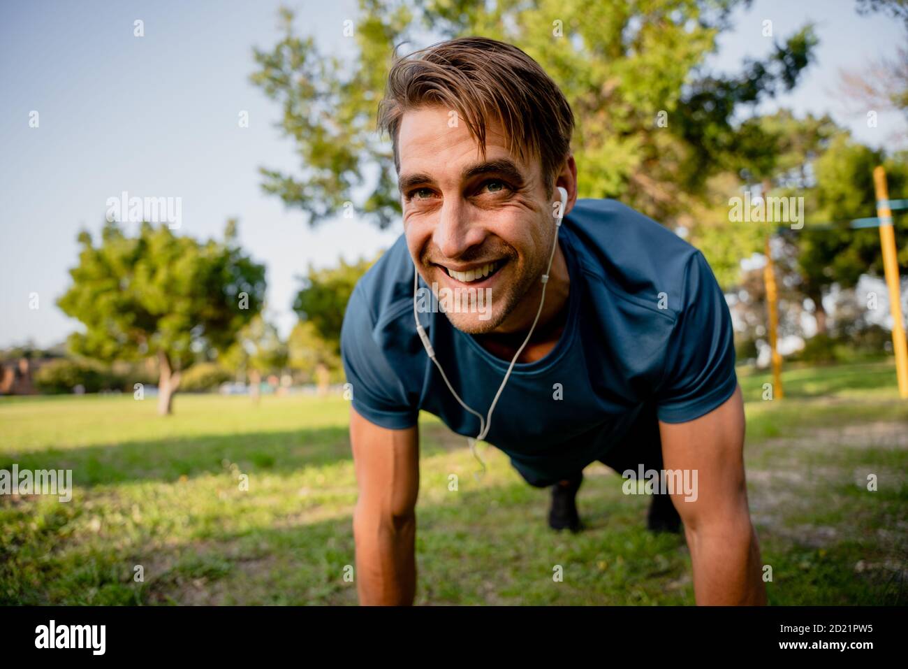 Sorridente bel giovane atleta maschile planking esercizio in soleggiata all'aperto i telefoni con le orecchie da parcheggio riproducono musica Foto Stock