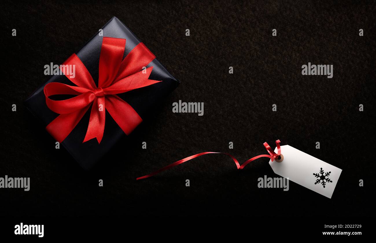 Un regalo di Natale avvolto in carta nera e nastro rosso con un'etichetta vuota, scheda su uno sfondo scuro dall'alto. Foto Stock