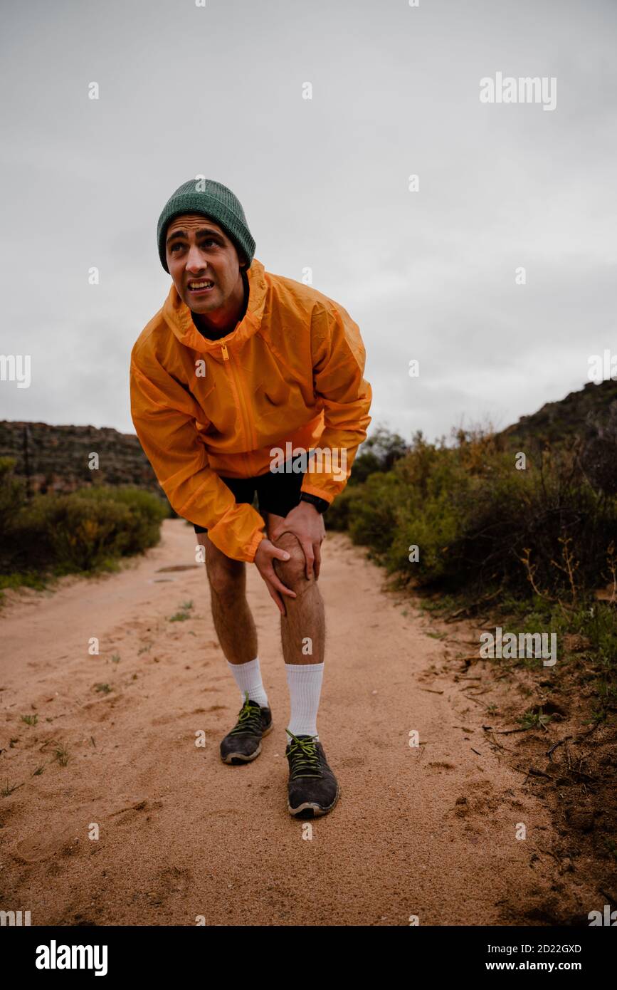 Sguardo doloroso su viso FIT giovane atleta maschile con ginocchio infortunio durante la corsa su sentiero di montagna bagnato in tempo nuvoloso Foto Stock
