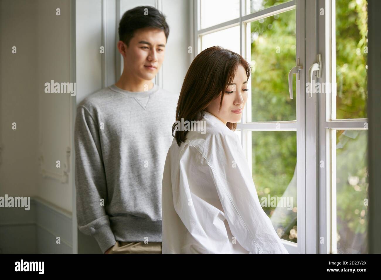 giovane coppia asiatica in piedi dalla finestra che parla chattando durante soggiorno a casa ordine Foto Stock