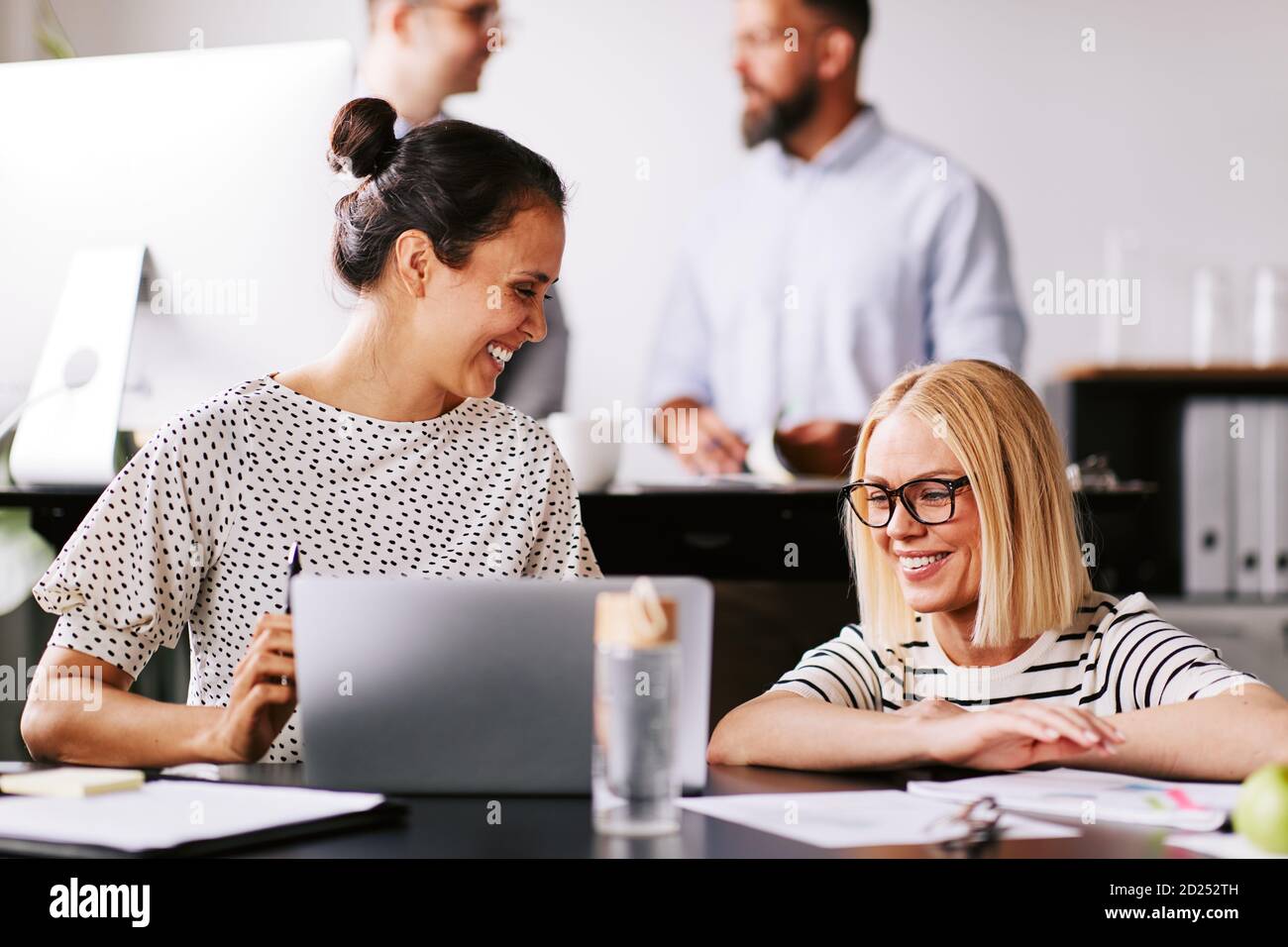 Due donne d'affari ridenti che parlano insieme su un computer portatile a un scrivania da ufficio con colleghi che lavorano in background Foto Stock