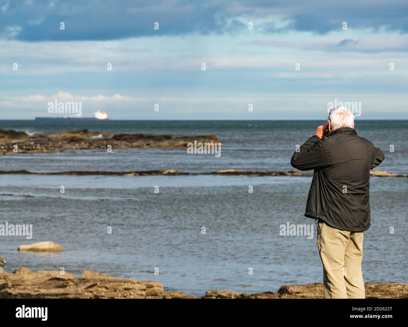 East Lothian, Scozia, Regno Unito, 6 ottobre 2020. Tempo in Gran Bretagna: Tempo in autunno a Ravensheugh Sands. Un uomo anziano guarda la vista attraverso il binocolo con una petroliera all'orizzonte Foto Stock