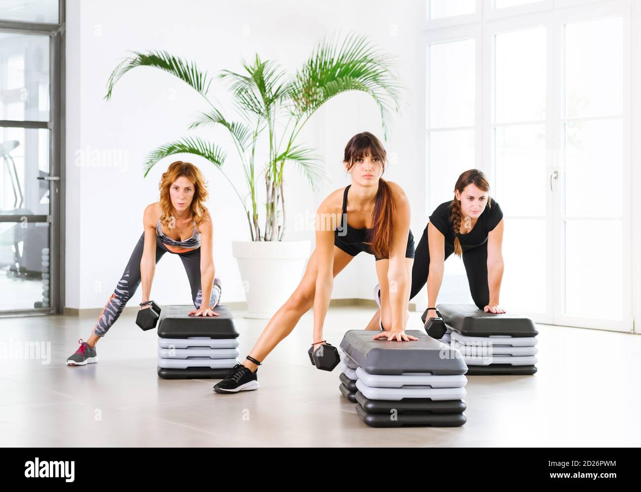 Tre giovani che si allenano in una classe di fitness utilizzando pesi dumbbell in una palestra ad alta chiave con piante in vaso in una salute e fitness o in una vita sana Foto Stock