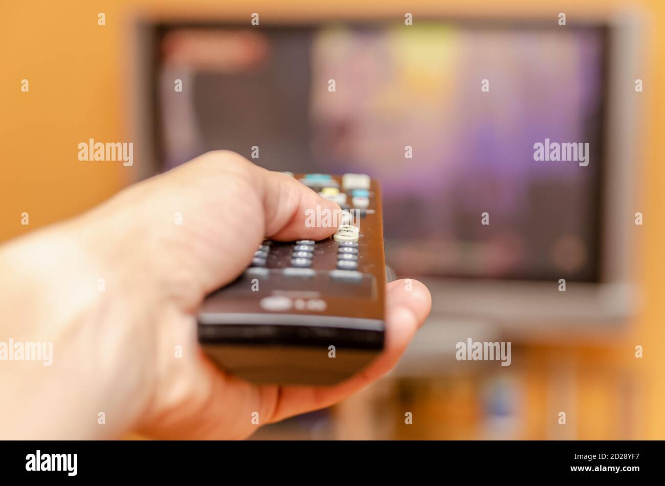 Mano dell'uomo che tiene un telecomando del televisore, premendo un pulsante mentre punta verso un televisore a schermo piatto. Foto Stock