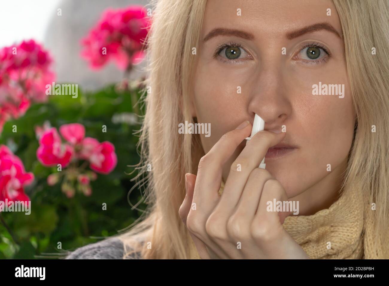 Giovane donna bionda usa uno spray nasale per un naso che cola o allergia contro uno sfondo di fiori interni, spazio di copia. Foto Stock