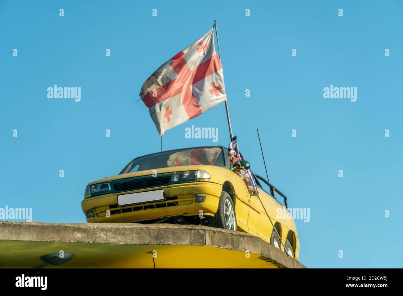 Auto gialla sul tetto dell'edificio con Bandiera georgiana contro il cielo blu Foto Stock