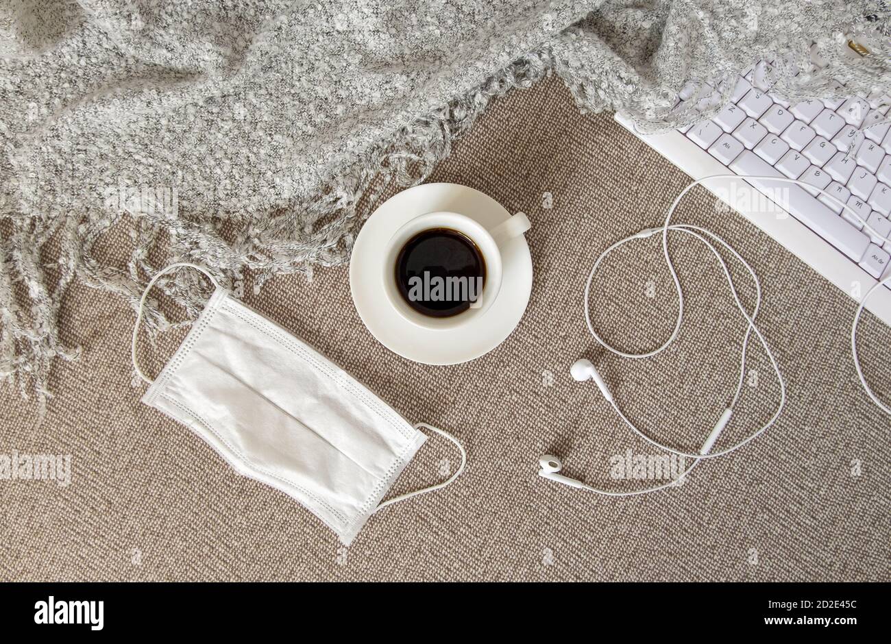 Una tazza di caffè, una tastiera, una cuffia, una plaid e una maschera medica sono sul divano. Foto Stock