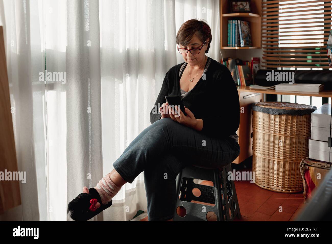 Donna adulta con capelli corti seduta con i pigiami utilizzando uno smartphone nella sua stanza studio mentre si trova a casa a causa della quarantena. Foto Stock