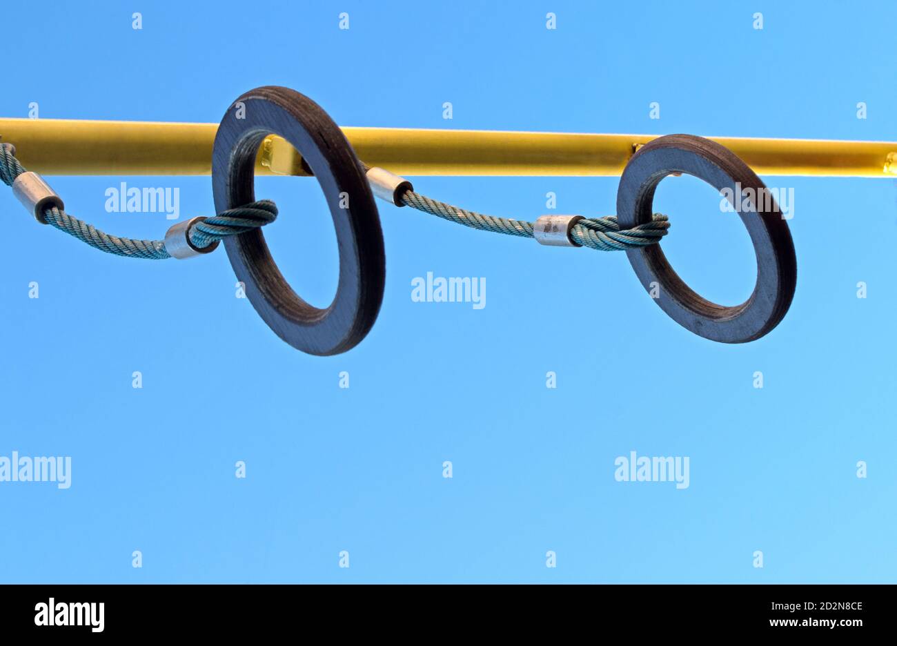 Gli anelli di gymnastic di legno marrone sono sospesi da corde blu ritorte con i cerchi di metallo compressi dalla traversa gialla sul parco giochi contro il cielo blu. Foto Stock