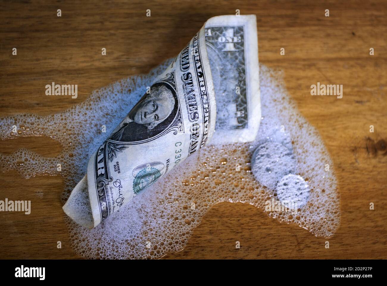 Una bolletta del dollaro arrotolata e monete, parzialmente ricoperte di schiuma su una superficie di legno Foto Stock