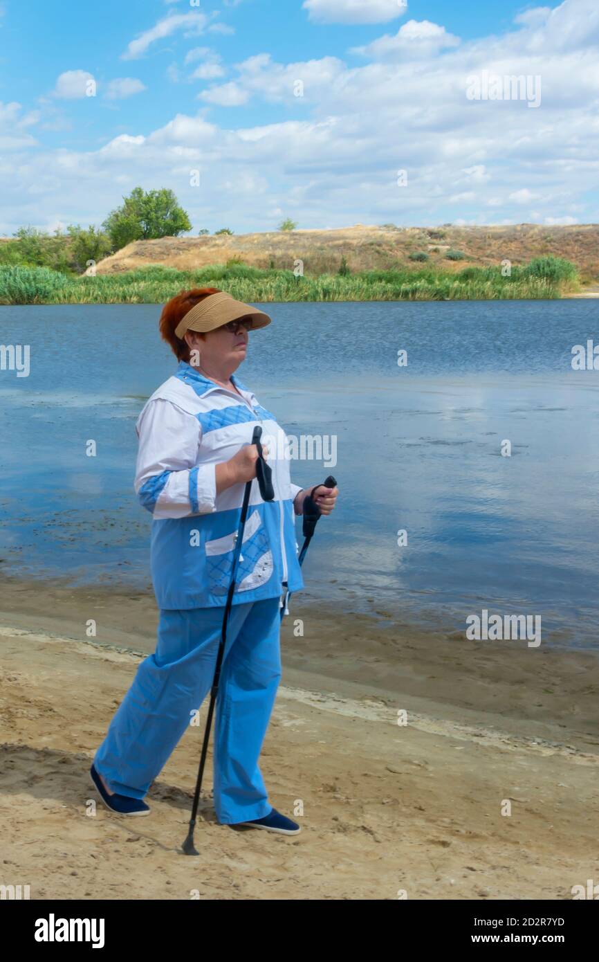 Donna anziana in tuta con bastoni nordici da passeggio sulla riva del fiume in estate. Concetto di stile di vita attivo nella vecchiaia. Messa a fuoco morbida, sfocatura. Foto Stock